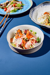 A vivid still life of an Asian salad with raw salmon, vegetables, and spicy sauce. Served in a bowl against a deep blue background with harsh shadows in an Asian restaurant setting. Vertical view.