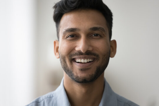 Cheerful handsome young Indian man front close up facial portrait. Face of happy attractive 20s guy with trendy stubble, black beard posing indoors, looking at camera with toothy smile