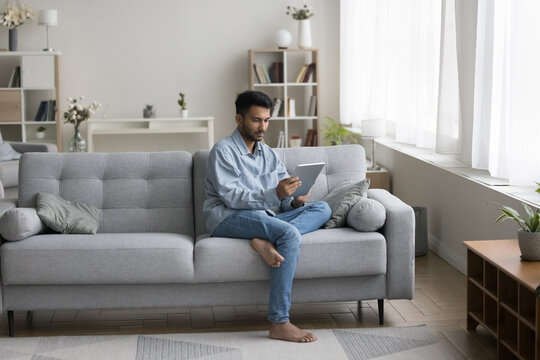 Focused Indian Student Guy Using Learning App On Tablet For Education, Internet Communication At Home, Holding Digital Computer, Relaxing On Couch, Looking At Electronic Screen. Full Length Shot