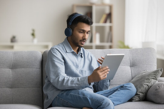 Serious young Indian man in headphones talking on video call at home, using tablet computer for online conference communication, conversation on Internet, speaking at screen