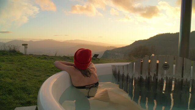 Zoom out drone shot of a young woman looking at camera and enjoying natural-fed hot tub. Breathtaking sunset in the outdoors. Outdoor bath is epic concept for spa weekend getaway or holiday retreat.