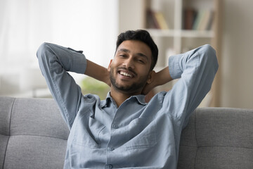 Happy attractive young Indian guy enjoying leisure, comfort at home, relaxing on comfortable sofa, stretching body, leaning on back, looking at camera with toothy smile. Video call head shot portrait