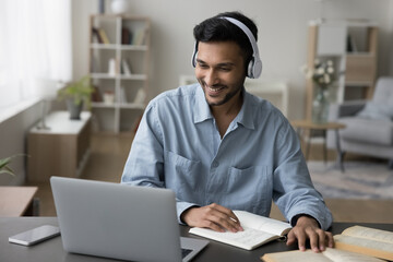 Cheerful young adult Indian student guy in headphones watching learning video on laptop, talking on video call to teacher, studying online, using Internet technology for education