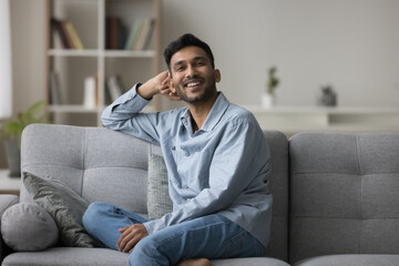 Cheerful handsome Indian guy relaxing on comfortable sofa at home, leaning hand on soft on back, enjoying comfort, leisure, looking at camera, smiling, laughing