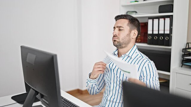 Young hispanic man business worker sweating using document as a hand fan at office