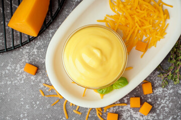 Plate with bowl of tasty cheddar sauce and grated cheese on black grunge background