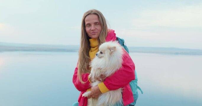 happy smiling female tourist holding white Spitz Volpino Italiano pet having rest on top of mountain, standing on hill enjoying beautiful landscape Slow motion friendship wondeful lake in background