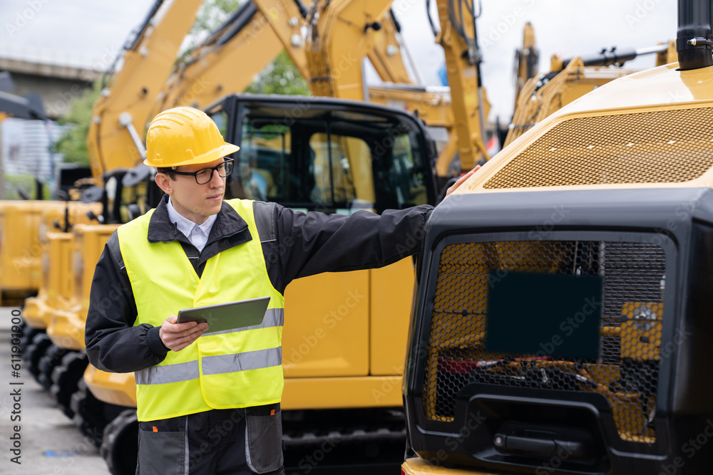 Poster engineer in a helmet with a digital tablet stands next to construction excavators