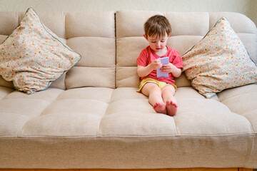 The content child is sitting on the couch with a phone in hand. The delighted kid lounges on the sofa, absorbed in their phone. Kid aged about two years (one year ten months)