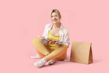Young woman with sushi and paper bag sitting on pink background