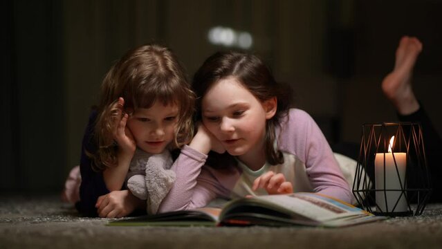 Two little girls are lying on a rug at home in the evening and reading a book