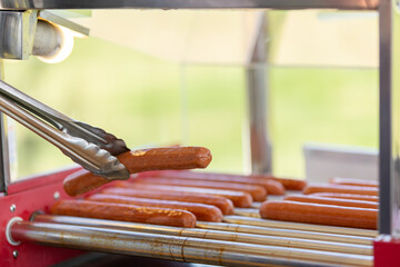Greasy hot dog sausages on a culinary machine in a cafe. Cooking sausages for a hot dog bun with sesame seeds in the background.