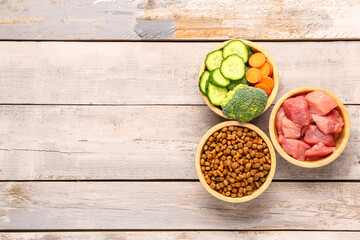 Bowls with dry pet food, raw meat and natural products on light wooden background