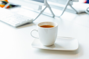 white cup with rectangular saucer with black coffee on white working table, workplace. cup, screen, keyboard on white table. total white composition. front view