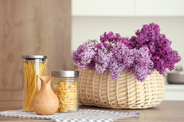 Basket with lilac flowers and jars of raw pasta on table in light kitchen