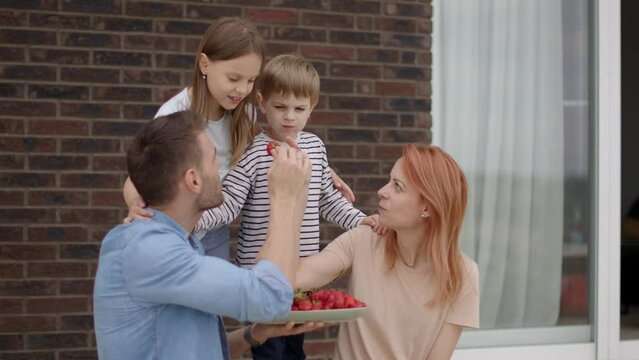 Family with a mother, father, son and daughter sitting outside on steps of a front porch of a brick house and eating strawberries