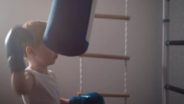 Focused little boy in boxer gloves punches hanging heavy bag in gym. Strong toddler develops new skills taking professional boxing classes slow motion