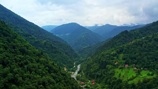 A river flowing between mountain ranges and aerial shots of village houses. Cloudy mountains and valleys. Aerial view of Kackar Mountains and Camlıhemsin. Green mountains and cloud covered sky. Turkey