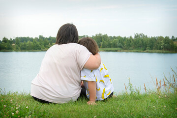 In summer, on the grass near the river, mother and overweight daughter sit embracing.