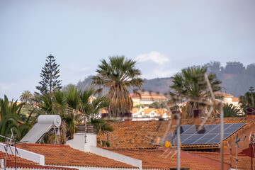 Solar panels on the roof of a single family house of red bricks and red tiles, with palm trees and landscape of trees in the background. Tenerife, Canary Islands, Spain