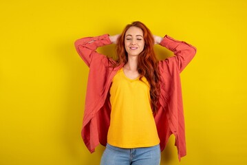 Close up portrait of Young redhead woman wearing fashion clothing over yellow wall who dreams about vacation holding hands behind head