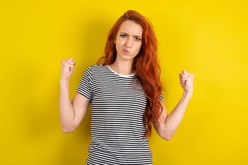 Irritated young redhead woman wearing  striped t-shirt over yellow wall blows cheeks with anger and...