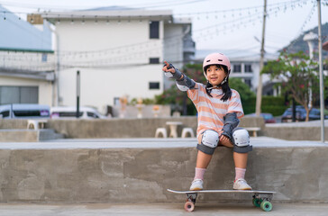 asian child skater or kid girl sitting relax at skatepark with skateboard or surf skate and pointing forward or people sports in street skate park to wears helmet elbow wrist knee guard by body safety