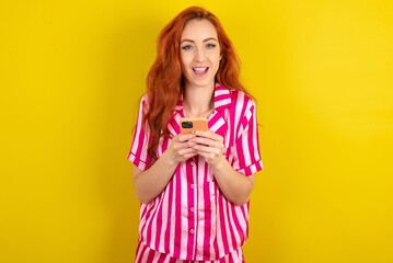 Excited Young red haired woman wearing pink pyjama over yellow studio background holding smartphone and looking amazed to the camera after receiving good news.