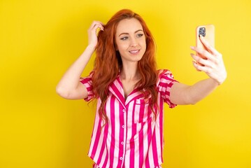 Young red haired woman wearing pink pyjama over yellow studio background smiling and taking a...