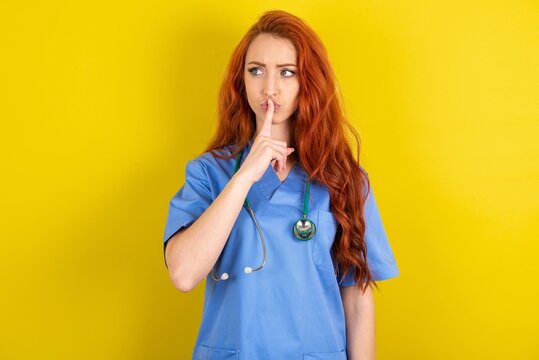young red-haired doctor woman over yellow studio background silence gesture keeps index finger to lips makes hush sign. Asks not to share secret