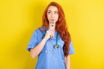 Photo of dreamy young red-haired doctor woman over yellow studio background lick fork look empty space - obrazy, fototapety, plakaty