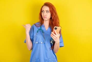 young red-haired doctor woman over yellow studio background points thumb away and shows blank space aside, holds mobile phone for sending text messages.