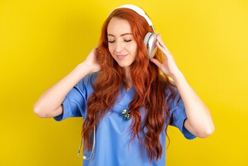 Pleased young red-haired doctor woman over yellow studio background enjoys listening pleasant melody keeps hands on stereo headphones closes eyes. Spending free time with music