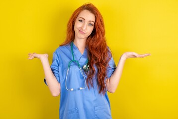 Careless attractive young red-haired doctor woman over yellow studio background shrugging shoulders, oops.