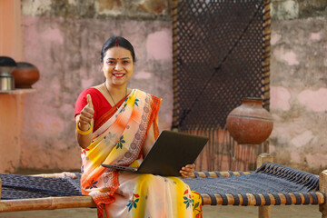 Happy rural Indian woman sitting outside the cottage holding a laptop and a card in her hands.