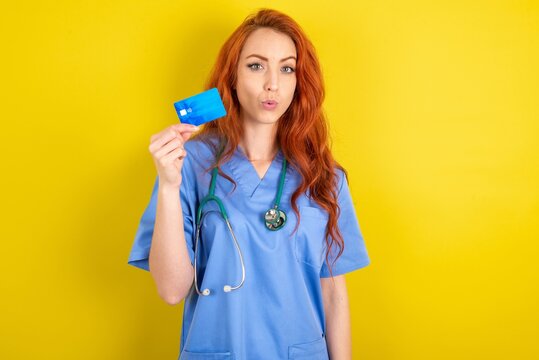 Photo of young red-haired doctor woman over yellow studio background amazed shocked hold credit card payment - Powered by Adobe