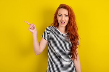 young beautiful red haired woman wearing striped t shirt over yellow studio background points aside on copy blank space. People promotion and advertising concept