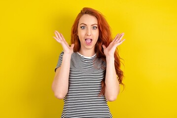 Surprised happy young beautiful red haired woman wearing striped t shirt over yellow studio...