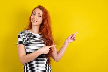 Optimistic young beautiful red haired woman wearing striped t shirt over yellow studio background points with both hands and  looking at empty space.