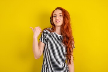 young beautiful red haired woman wearing striped shirt over yellow studio background listens audio...