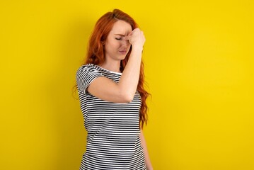 Sad young beautiful red haired woman wearing striped shirt over yellow studio background suffering from headache holding hand on her face