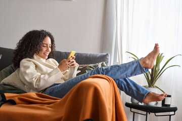 Smiling relaxed young latin woman sitting on couch using cell phone at home. Happy barefoot girl holding smartphone, looking at cellphone doing shopping in mobile digital apps buying online on sofa.