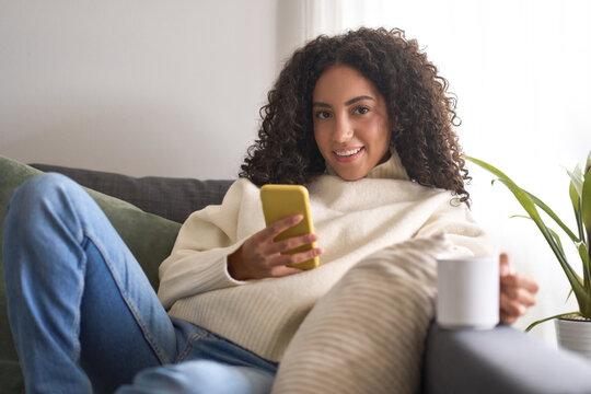 Young pretty latin woman sitting on couch using cell phone, holding smartphone looking at camera wit cellphone doing online shopping in mobile digital shop app, checking social media commercial offer.