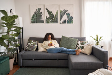Relaxed latin girl sitting on couch using mobile cell phone at home. Calm young woman relaxing on...