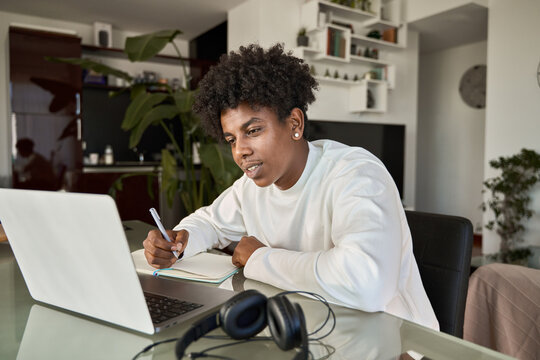 African American student elearning online class writing notes at home. Teenage boy using computer watching webinar, learning virtual lesson, taking hybrid web course writing notes sitting at table.