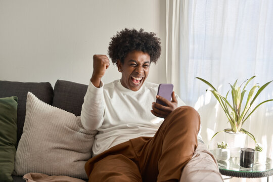 Happy excited gen z African American guy sitting on couch at home holding cellphone, celebrating online digital money betting win with yes gesture using mobile app on cell phone. Authentic shot.