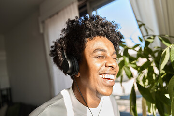 Happy funky gen z hipster African American guy wearing headphones laughing at home, listening music, having fun feeling relaxed standing in living room at home. Close up authentic shot. - obrazy, fototapety, plakaty