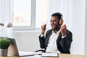 An African-American man sits at his desk in front of his laptop, wearing headphones and chatting on a video call, listening to music. The concept of student business training and online work.