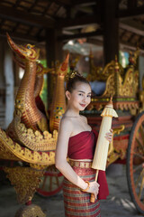 Pretty Asian girl wearing ancient native Northern Thai traditional dress costumes posing and holding the umbrella with a Siam royal Chariot car in the ceremony background.