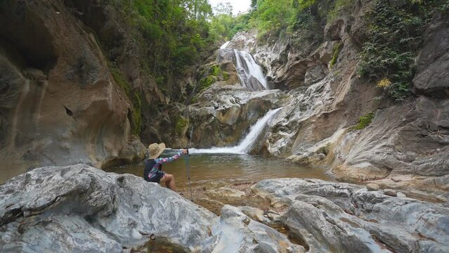 asian child camper or kid girl trekking hiking by walking pole and wearing hat to sitting relax on rocks and watching nature waterfall in forest for holiday adventure travel at lan sang national park